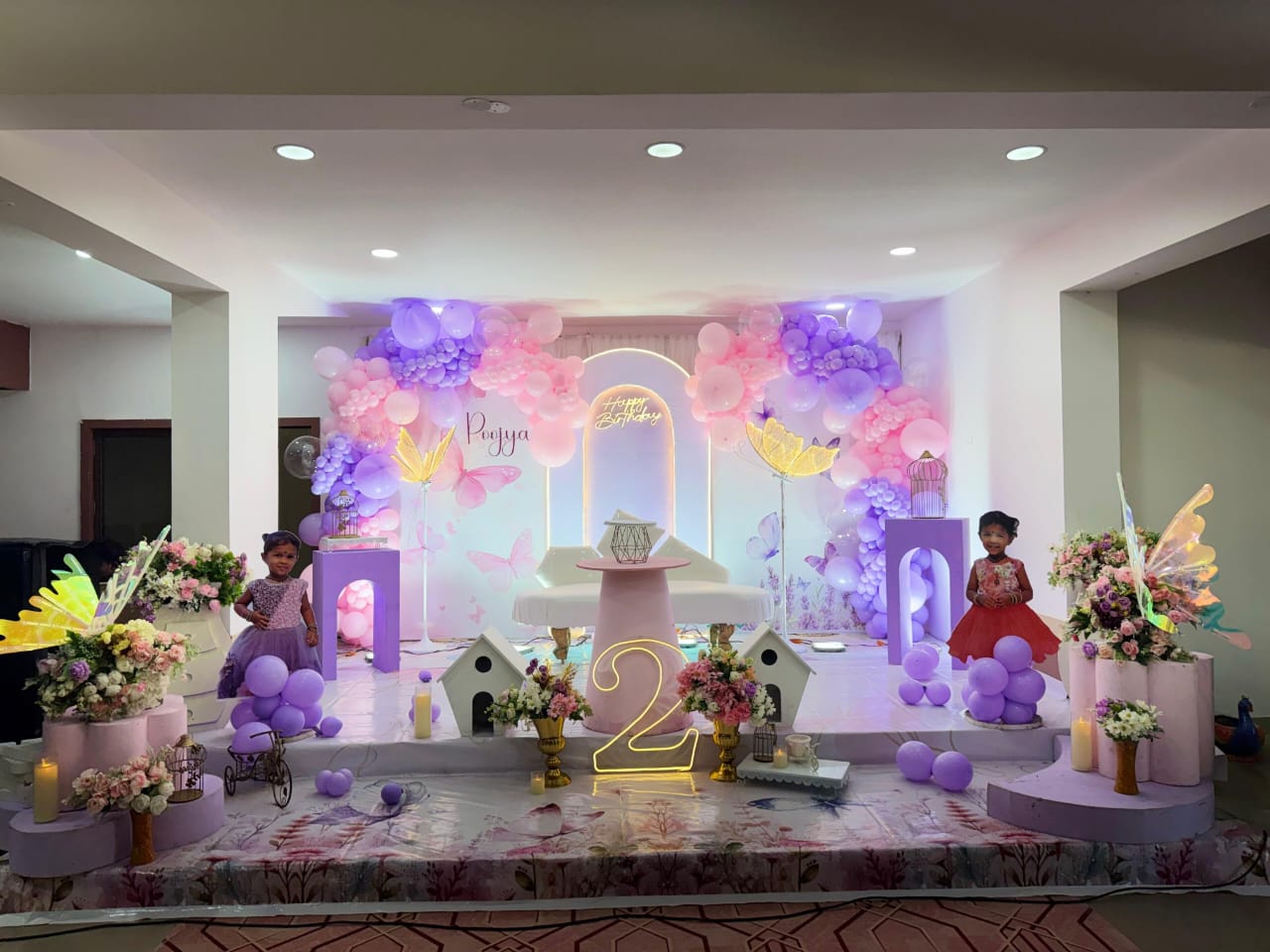 birthday celebration stage with pastel balloon arch, butterflies and neon sign